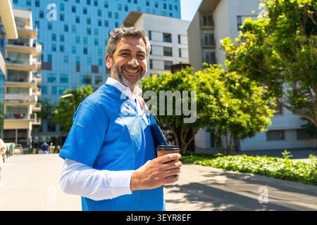 Doktor lächelt in Peelings und hält Kaffee vor dem Krankenhaus Stockfoto