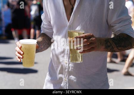 Mann mit Tattoos hält zwei Plastikbecher kaltes Bier und genießt die sonnige Atmosphäre beim lebendigen Southsea Food Festival in Portsmouth, England Stockfoto