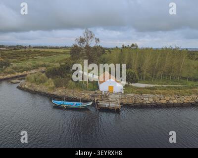 Blick aus der Vogelperspektive auf die Aveiro Lagune mit einem kleinen weißen Gebäude, einem hölzernen Dock und einem blauen Boot, das auf dem Wasser im Aveiro District, Portugal, verankert ist. Stockfoto
