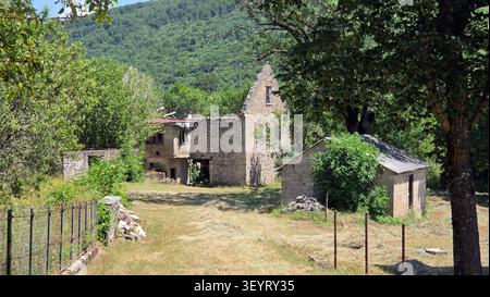 Ein altes Steinhaus liegt verfallen und zerbröckeln auf einem grasbewachsenen Feld, umgeben von üppigen Bäumen und einer steilen Berglandschaft Stockfoto
