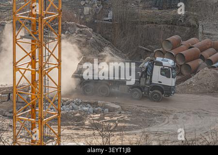 Baustelle mit großem Kipplaster, der sich durch staubige Wege bewegt und Industriemaschinen und laufende Arbeiten hervorhebt, mit einem hohen orangefarbenen Met Stockfoto
