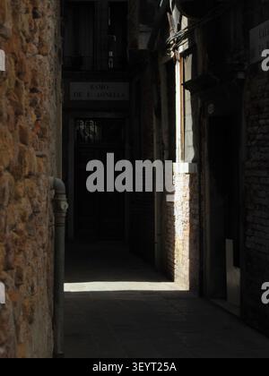 Dunkle, enge Gasse in Venedig, Italien, mit einem Schacht aus Sonnenlicht, der die Ziegelmauer beleuchtet Stockfoto