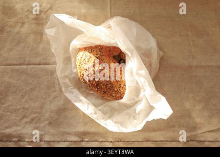 Eine Nahaufnahme eines gerösteten Sesambagels, verpackt in einer weißen Papiertüte auf einer beigen texturierten Textoberfläche. Stockfoto