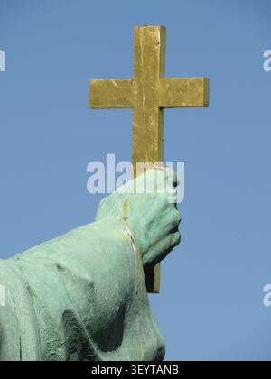 Eine Nahaufnahme der Hand einer Bronzestatue, die ein goldenes Kreuz mit einem blauen Himmel im Hintergrund hält. Die Statue ist grün und der Himmel blau. Stockfoto