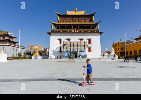 Ein Kind, das auf einem Roller vor dem Kloster Gandantegchinlen fährt, einem buddhistischen Tempel in Ulaanbaatar, Mongolei, wo Tradition und modernes Leben verschmelzen. Stockfoto