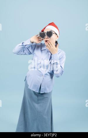 Schwangere Frau mit Ferienhut mit Sonnenbrille im Studio mit Blau Stockfoto