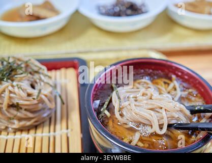 Gekühlte Buchweizennudeln auf einer Bambusmatte mit herzhafter Dip-Sauce. Essstäbchen heben einen Bissen, der eine erfrischende authentische asiatische Sommer Mahlzeit einfängt. Stockfoto