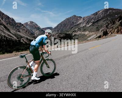 Ein männlicher Radfahrer erklimmt eine weit geschwungene Bergpassstraße auf einem bronzenen Rennrad unter dramatischen felsigen Sierra-Nevada-Gipfeln an einem klaren Sonnentag. Stockfoto