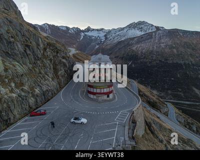 Blick aus der Vogelperspektive auf das Hotel Belvedere an der berühmten Haarnadelkurve des Furka-Passes, mit zwei geparkten Sportwagen, umgeben von zerklüfteten Bergen und sn Stockfoto