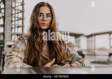 Frau Brille Smartphone sitzt an einem Tisch im Freien und trägt eine stilvolle Brille für ein lässiges Lifestyle-Porträt Stockfoto