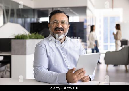 Glücklicher, reifer indischer Geschäftsmann, der ein digitales Tablet am Schreibtisch hält Stockfoto