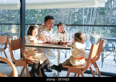 Die Familie versammelt sich um den Tisch und lächelt warm. Kinder greifen zueinander, voller Neugier. Weiches Licht leuchtet durch große Fenster hinter ihnen. Stockfoto