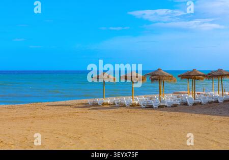 Reihen von Strohschirmen und weißen Sonnenliegen säumen einen unberührten Sandstrand, umgeben von türkisfarbenem Mittelmeer-Wasser unter einer Wolke Stockfoto