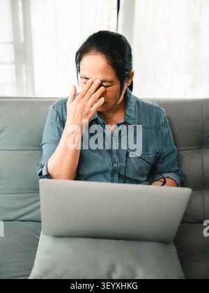 Müde asiatische Frau reibt sich die Augen, während sie zu Hause mit dem Laptop arbeitet, eine Brille hält, das Konzept der Augenbeanspruchung, digitale Ermüdung, Kopfschmerzen, Bildschirmzeit und mehr Stockfoto