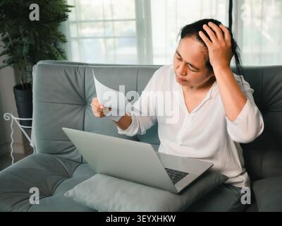 Besorgte asiatische Frau mittleren Alters, die Papierdokumente neben dem Laptop zu Hause liest und den Kopf mit Stress über Rechnungen, finanzielle Probleme, Papierkram oder berührt Stockfoto