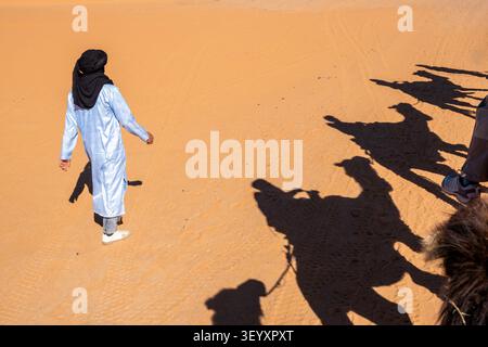 Hohe Blickwinkelaufnahme eines marokkanischen Kameltreibers in einem blauen Gewand, der an einem sonnigen Tag in Merzou auf Sand neben den langen dunklen Schatten einer Kamelkarawane geht Stockfoto