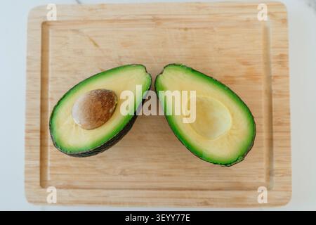 Eine frische Avocado, die in zwei Hälften geschnitten und auf ein hölzernes Schneidebrett gelegt wird. Perfekter kulinarischer Hintergrund für Rezepte, Diäten zur Gewichtsreduktion und sauberes Essen. Oben Stockfoto