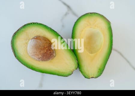 Blick von oben auf eine frische reife Avocado, die in zwei Hälften geschnitten wurde, auf einem sauberen weißen Marmorhintergrund. Eine Hälfte enthält die Grube, die ein gesundes Esskonzept darstellt. Schließen Stockfoto