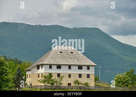 Ein altes ethnographisches Museum aus Stein verfügt über ein Schindeldach und weiße Wände, umgeben von einem Holzzaun vor einer Berglandschaft. Stockfoto