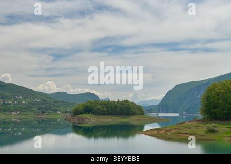 Üppig grüne Insel mitten im Rama Lake, mit ruhigen Reflexen, umliegenden Bergen und friedlicher Natur unter einem bewölkten Himmel. Stockfoto