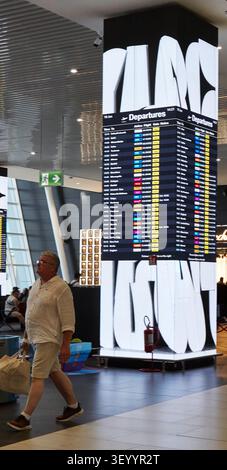 Fiumicino, Italien - 19. Juli 2026: Auf der Anzeigetafel zum Flughafenterminal werden die Abflüge aufgelistet, während ein Mann in kurzen Hosen mit einem Shoppin spaziert Stockfoto