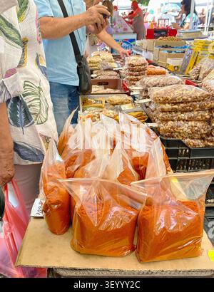 Abgepackte Gewürze und Snacks werden auf einem Tisch auf einem ländlichen Markt serviert, bei dem die Käufer interagieren und verschiedene Essensoptionen im Hintergrund erkunden Stockfoto