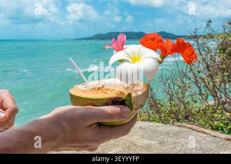 Frisches Kokos-Eis mit tropischen Blüten in männlichen Händen. Tropisches Sommerdessert mit türkisfarbenem Wasser im Hintergrund, Samui Island, Thailand Stockfoto