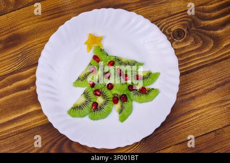 Weihnachtsbaum aus Kiwischeiben und Granatapfel auf Holztisch. Draufsicht. Kreative Idee für festliche Weihnachts- und Neujahrsdesserts. Lustige Lebensmittel-ID Stockfoto