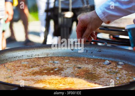 Der Koch fügt eine große Metallpfanne mit Paella im Freien mit verschwommenen Personen und Kochgeräten im Hintergrund hinzu, was ein lebendiges Kochen schafft Stockfoto