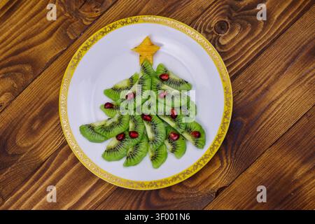 Weihnachtsbaum aus Kiwischeiben und Granatapfel auf Holztisch. Draufsicht. Kreative Idee für festliche Weihnachts- und Neujahrsdesserts. Lustige Lebensmittel-ID Stockfoto
