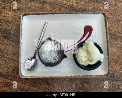 Geschmolzener Schokoladenkuchen, begleitet von einer Schaufel Vanilleeis und Himbeersauce auf einem rechteckigen Teller mit einem Löffel daneben Stockfoto