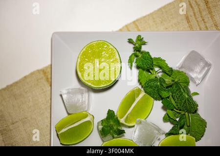 Nahaufnahme von oben nach unten mit frischen grünen Limettenkeilen, organischen Minzblättern und klaren Eiswürfeln auf einer weißen Keramikplatte. Rustikale Sackleinen Stockfoto