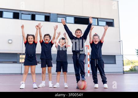 Gruppe aufgeregter Kinder in Uniform, die mit erhobenen Armen jubeln und nach dem Spielen auf einem Sportplatz stehen Stockfoto