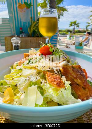 Caesar-Salat mit gegrilltem Huhn und Parmesankäse, serviert mit einem Glas Bier in einem Strandrestaurant im Freien Stockfoto