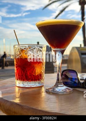 Espresso-Martini- und negroni-Cocktails auf einem Holztisch an einer Outdoor-Marina-Bar bei Sonnenuntergang Stockfoto