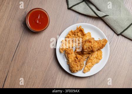 Knusprige Hähnchenflügel und Streifen auf einem weißen Teller werden mit roter Dippsoße und einer gefalteten Leinenserviette auf einer Holzoberfläche kombiniert. Perfekt für Essen-BHs Stockfoto