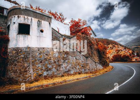 Weitwinkelblick auf ein verwittertes leeres Gebäude mit Steinmauer, geschwungener Bergstraße, rotem Herbstlaub und dramatischem bewölktem Himmel Stockfoto