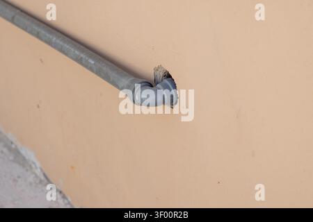 Eine Nahansicht zeigt ein graues PVC-Rohr mit einem 90-Grad-Winkelstück, das in einer gezackten Öffnung in einer strukturierten pfirsichfarbenen Wand mit Betonvisibl verschwindet Stockfoto