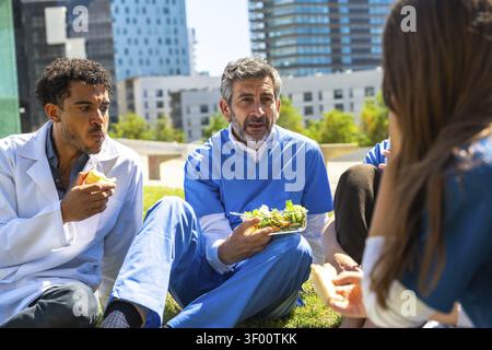 Medizinische Fachkräfte entspannen und genießen eine gesunde Mahlzeit auf dem Gras außerhalb des Krankenhauses, fördern Wohlbefinden, Teamarbeit und ein ausgewogenes Lifest Stockfoto