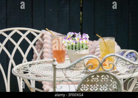 Hausgemachte Rosa- und Zitruslimonade, serviert mit frischem Gebäck auf einem eleganten schmiedeeisernen Gartentisch, dekoriert mit blühenden Stiefmütterchen. Stockfoto