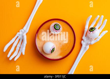 Eine Schüssel Kürbissuppe mit verspielten Augapfeldekorationen, perfekt für ein kreatives Halloween-Vergnügen. Vor einem leuchtenden orangefarbenen Hintergrund Stockfoto