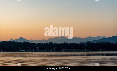 Pastellfarbener Sonnenuntergangshimmel über dem Genfer See, mit ruhigem Wasser, Hafenmasten und Stadtlichtern im Vordergrund und dem Mont Blanc-Massiv und den französischen Alpen in Silh Stockfoto