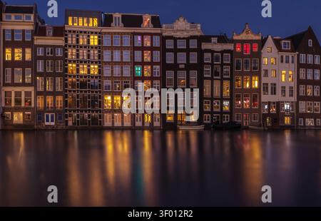 Nächtlicher Blick auf Amsterdams berühmte Tanzhäuser entlang eines Kanals, glühende Fenster und warme Lichter, die sich auf ruhigem Wasser spiegeln, schaffen ein bezauberndes HIS Stockfoto