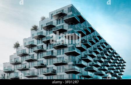 Moderner, abgestufter Apartmentblock mit sich wiederholenden Glasbalkonen und geometrischer Fassade vor einem bewölkten Himmel, der moderne urbane Häuserarchitektur zeigt Stockfoto