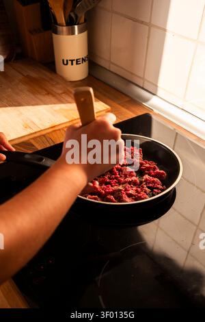 Nahaufnahme der Hände, die Hackfleisch in einer Pfanne auf einem eleganten Induktionskochfeld rühren, mit warmem Sonnenlicht und Küchenutensilien im Hintergrund, Zubereitung Stockfoto