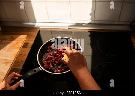 Nahaufnahme von Händen, die Hackfleisch in einer Pfanne auf einem schwarzen Glaskochfeld rühren, mit warmem Sonnenlicht und gefliestem Backsplash, die Hausmannskost und vieles mehr einfangen Stockfoto
