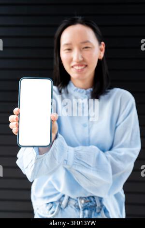Gen Z asiatische Frau lächelt und hält ein Smartphone mit einem leeren weißen Bildschirm für ein Modell. Lässiges Outfit bei weichem Tageslicht vor dunklem Hintergrund, ide Stockfoto