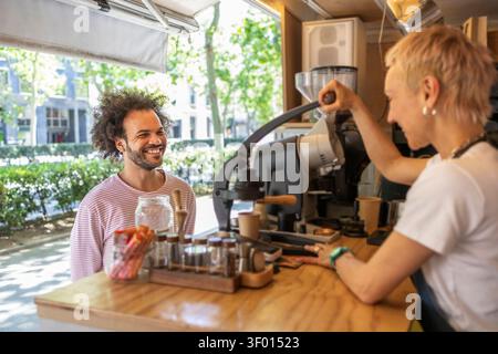 Ein freundlicher Barista serviert einem lächelnden Gast Kaffee zum Mitnehmen an einem Kaffeekiosk für kleine Unternehmen mit Espresso-Maschine und Mahlwerk in einer hellen Straße Stockfoto