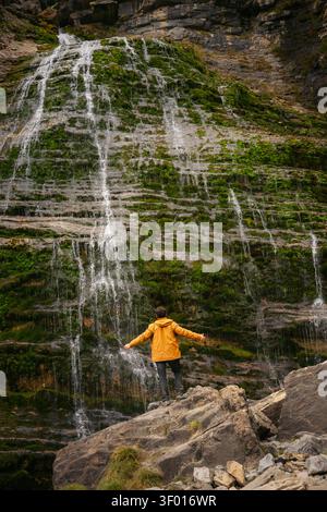 Ein einsamer Wanderer in gelber Jacke steht auf felsigem Boden unter dem Horsetail Waterfall, mit dünnen Bächen, die über eine moosige Klippe in Ordesa und Monte P kaskadieren Stockfoto