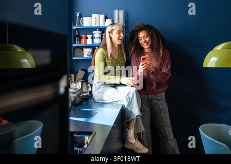 Zwei multiethnische Mitbewohner lachen, während sie in einer stilvollen Küche auf ein Smartphone schauen, sich zu Hause entspannen und Freundschaft genießen, zwangloses Leben und Spaß haben Stockfoto